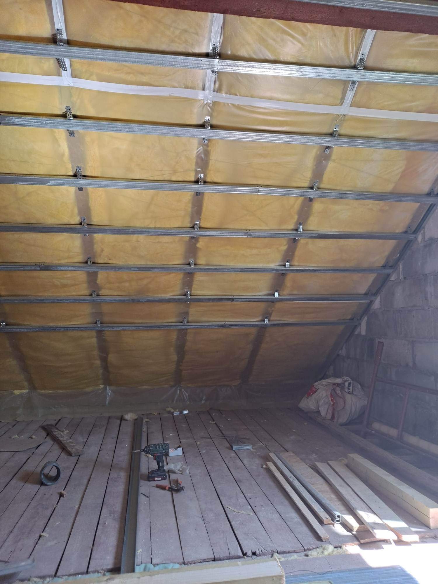 Poddasze w trakcie ocieplania: widoczne metalowe profile konstrukcyjne, warstwa izolacji z folią, drewniana podłoga, narzędzia i materiały budowlane porozrzucane na podłodze.