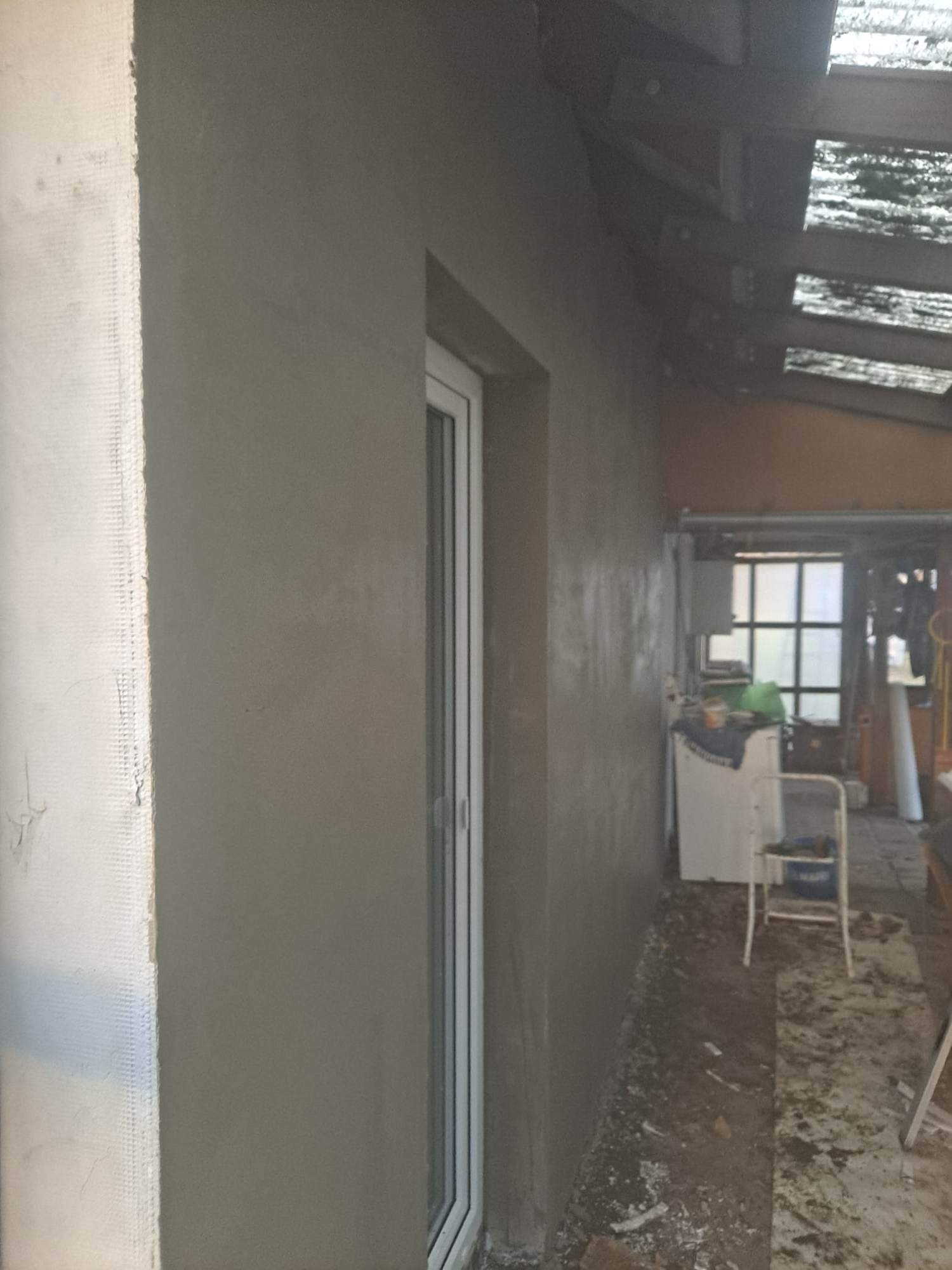 Świeżo otynkowana ściana zewnętrzna budynku z zamontowanym oknem PCV, widoczne siatka zbrojeniowa na narożniku i niedoskonałości tynku w pomieszczeniu gospodarczym.