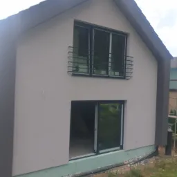 Dwukondygnacyjny dom z jasnoszarą elewacją, czarnymi ramami okiennymi i balkonem z metalową balustradą. Widoczny fragment zielonej izolacji przy gruncie i fragment rynny.