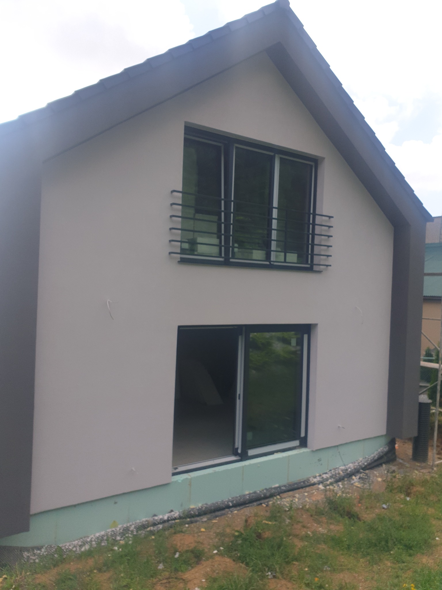 Dwukondygnacyjny dom jednorodzinny z jasnoszarą elewacją i czarnymi ramami okiennymi, widok z zewnątrz na niezabudowany parter i balkon z metalową balustradą na piętrze.