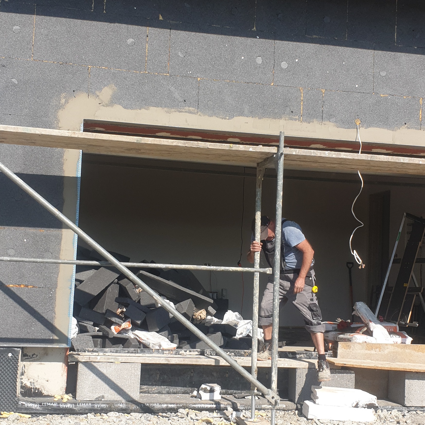 Pracownik na rusztowaniu przy ocieplonej ścianie budynku, widoczne płyty styropianowe i fragmenty szarej elewacji, przygotowane materiały izolacyjne w otworze okiennym.