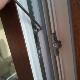 Regulacja okna PCV za pomocą klucza imbusowego, widoczny mechanizm blokujący i fragment ramy okiennej w kolorze drewna.