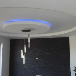 Dekoracyjny sufit podwieszany z oświetleniem LED i nowoczesną lampą wiszącą w jasnym wnętrzu z ciemną ścianą z kamienia.