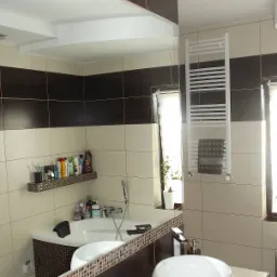 Elegancka łazienka z beżowo-brązowymi płytkami, owalną umywalką i grzejnikiem drabinkowym. Widoczne lustro odbija wannę z mozaikową obudową i półką z kosmetykami.