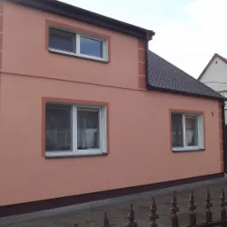 Dwukondygnacyjny dom z elewacją w odcieniach brzoskwini, z brązowymi akcentami wokół okien i narożników. Widoczne białe okna z firankami i brązowy dach.