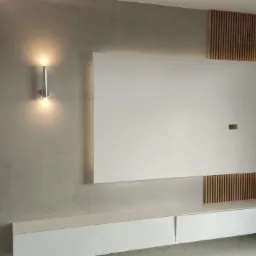 Ściana w salonie z szarą, betonową fakturą, podświetlona kinkietem, z panelem TV otoczonym drewnianymi lamelami i białą szafką RTV na dole.