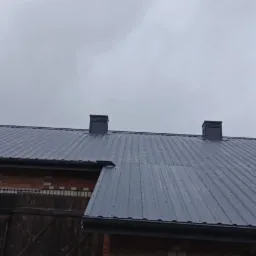 Szary dach z blachy trapezowej na budynku z czerwonej cegły, widoczne dwa kominy i rynna, pochmurne niebo w tle.