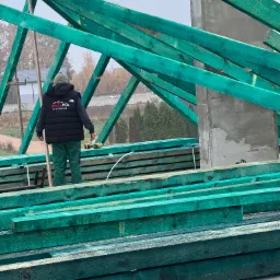 Monter w czarnej kamizelce firmy Dach-Pol montuje zieloną więźbę dachową; w tle komin i zabudowa jednorodzinna.