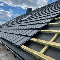 Montaż czarnej blachodachówki modułowej na drewnianej konstrukcji z widoczną membraną dachową, rynna w dolnej części kadru, błękitne niebo z chmurami w tle.