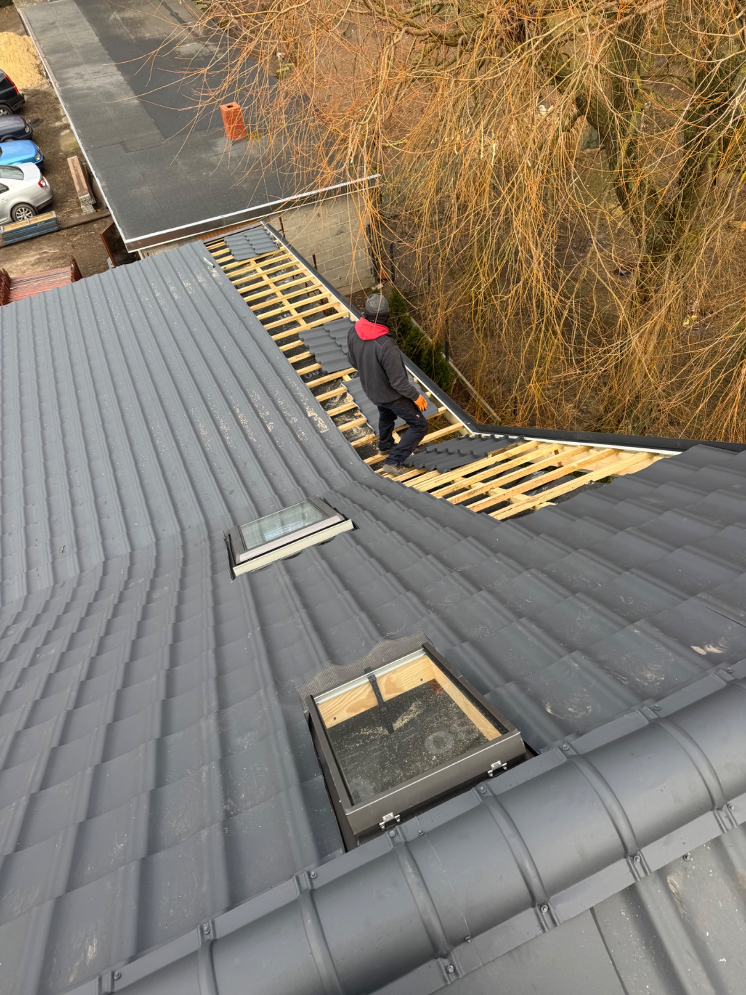 Pracownik w pomarańczowych rękawicach montuje szare panele dachowe na drewnianej konstrukcji z widocznymi oknami dachowymi, z perspektywy z góry.