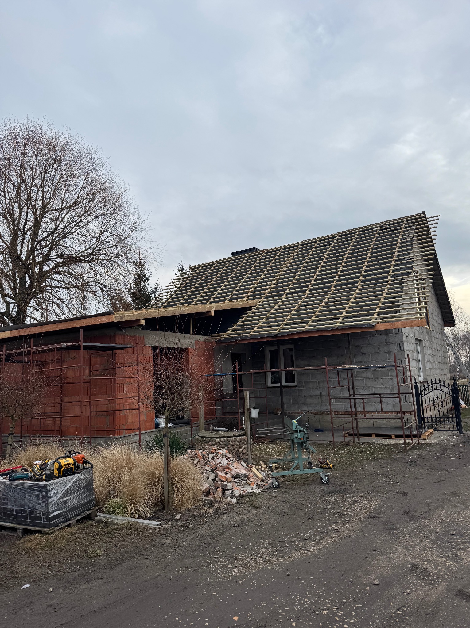 Budynek w trakcie remontu dachu; widoczna konstrukcja więźby dachowej, rusztowania, materiały budowlane i narzędzia na ziemi.