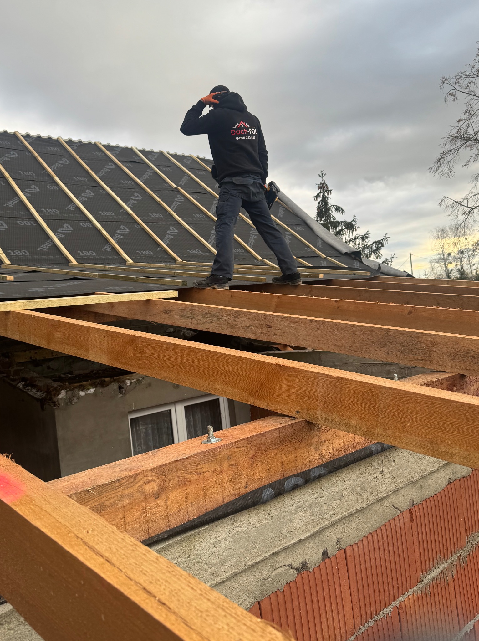 Monter w czarnej bluzie z logo firmy na więźbie dachowej, zabezpieczonej membraną, w trakcie prac dekarskich na tle pochmurnego nieba.
