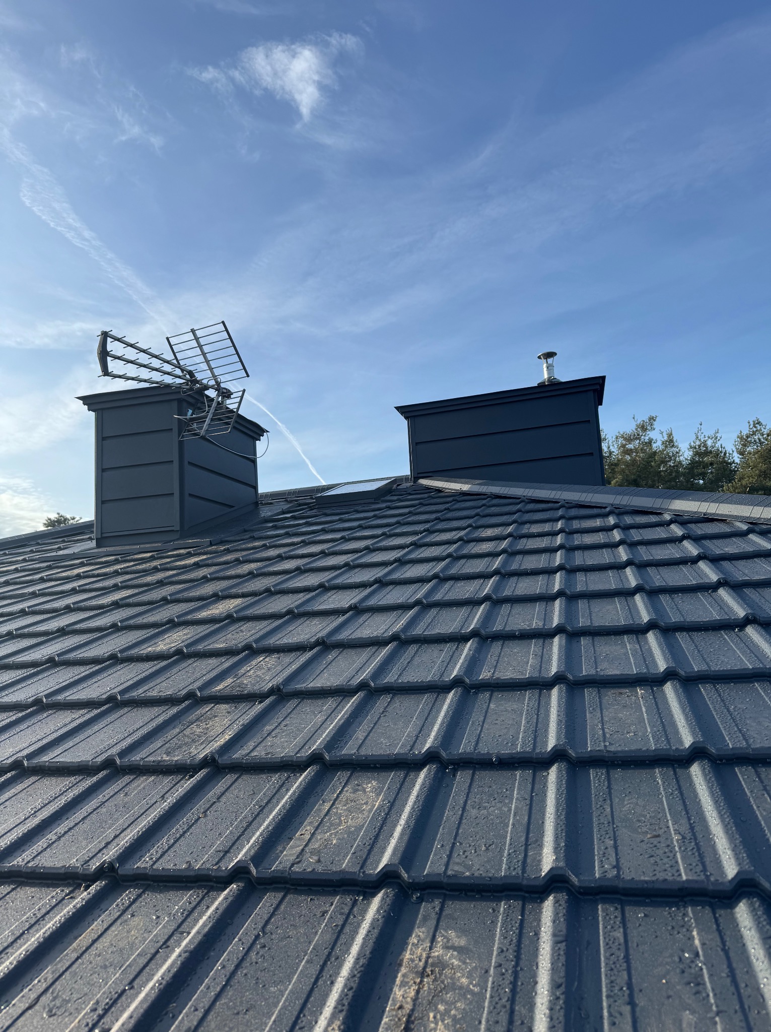 Grafitowy dach z dachówki modułowej, dwa kominy z obróbkami blacharskimi, antena telewizyjna, błękitne niebo z smugami kondensacyjnymi.