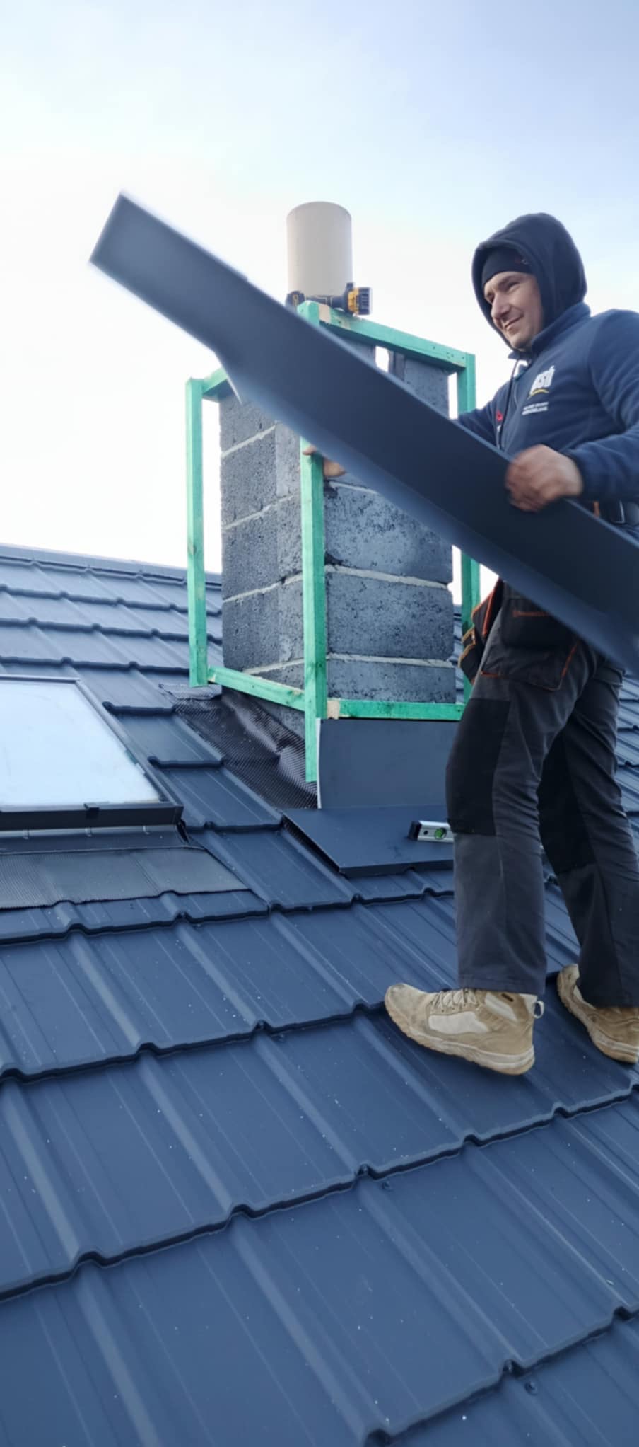 Dekarz na dachu z blachy trapezowej montuje obróbkę komina z użyciem drewnianej konstrukcji pomocniczej.