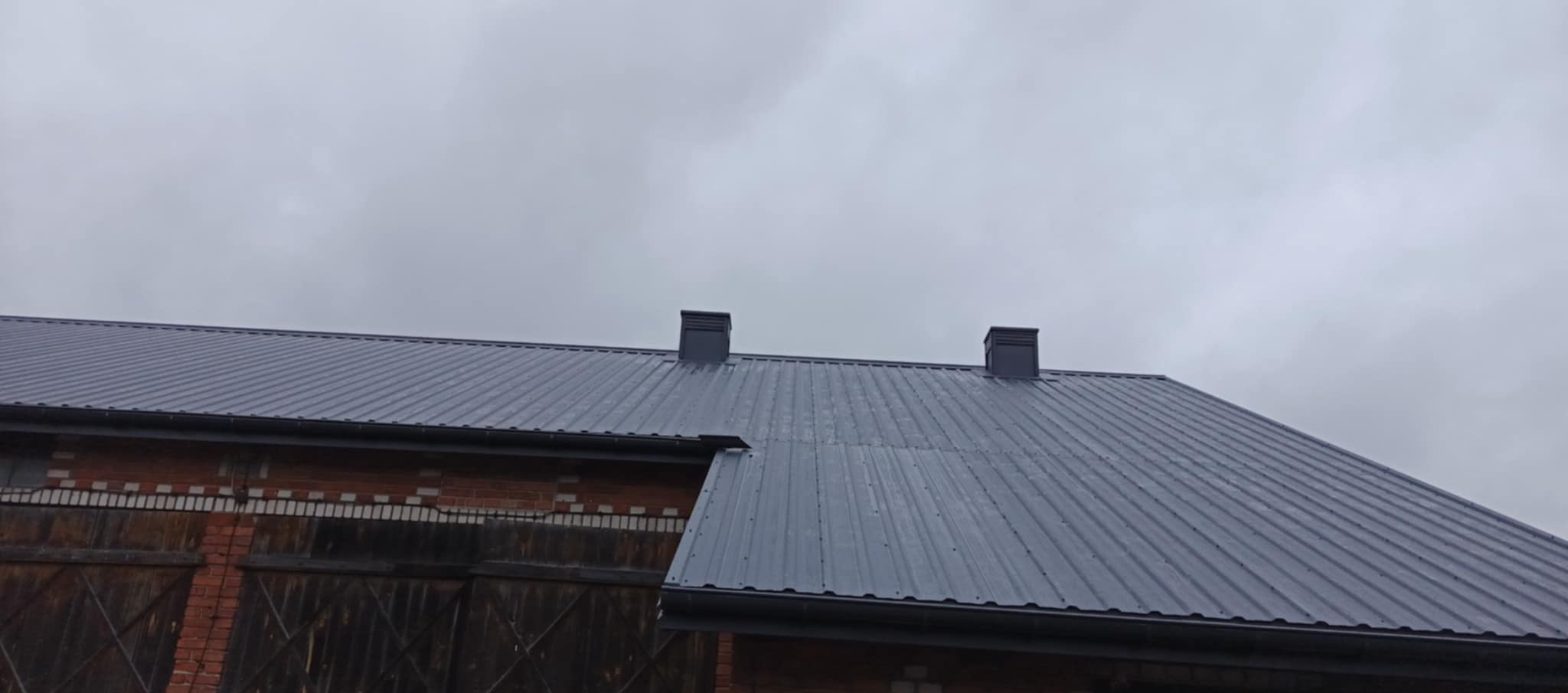Szary dach z blachy trapezowej na budynku z czerwonej cegły, widoczne dwa kominy i rynna, pochmurne niebo w tle.