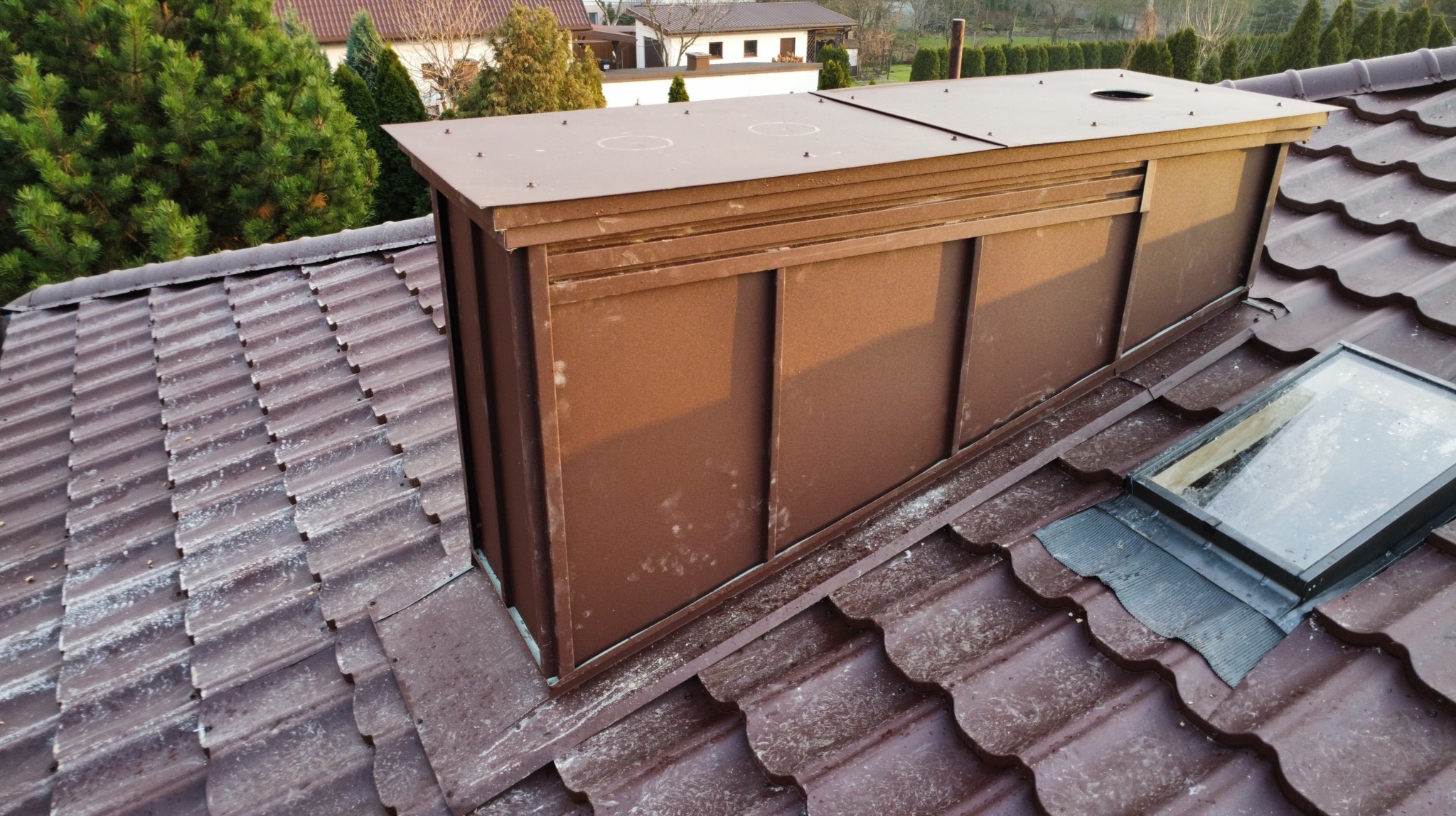 Brązowy komin wentylacyjny z blachy na dachu z dachówki ceramicznej, widoczne okno dachowe i otoczenie z zielenią.