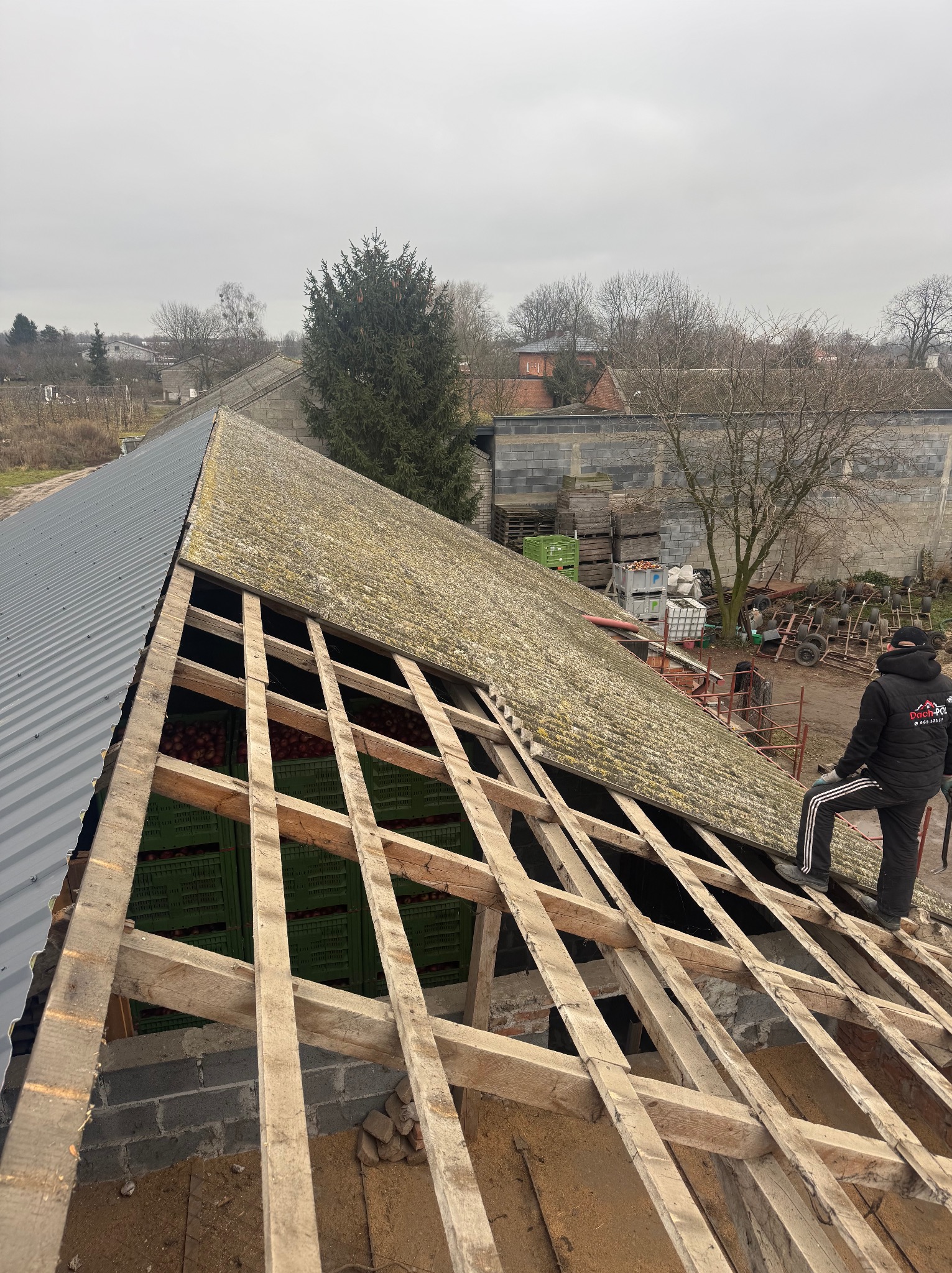 Demontaż starego dachu z eternitu, widoczna drewniana konstrukcja więźby oraz nowa blacha trapezowa na części dachu, w tle budynki gospodarcze i pracownik w kurtce firmowej.
