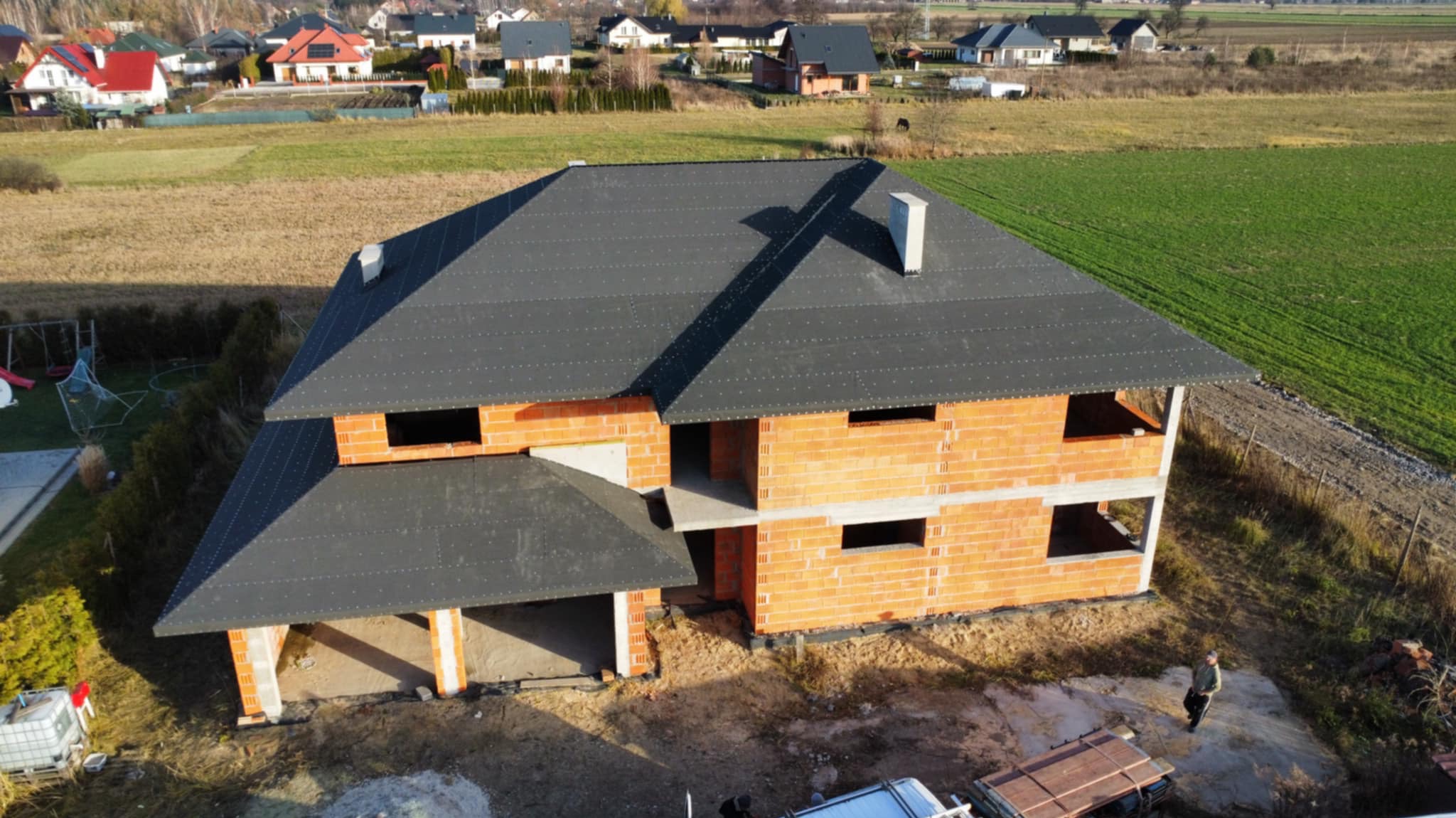 Dom w trakcie budowy z nowym, ciemnoszarym dachem, widok z góry; murarz stoi obok budynku.