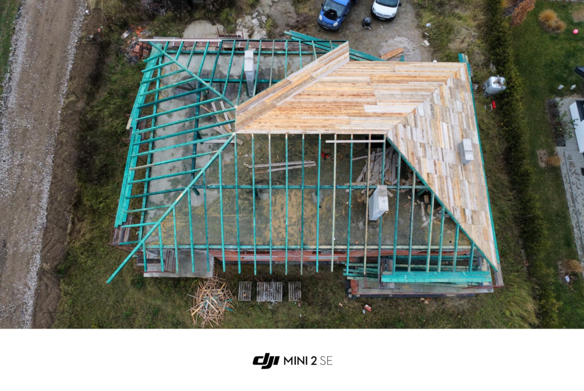 Widok z lotu ptaka na budowę dachu z częściowo ułożonymi drewnianymi deskami i pomalowaną na zielono konstrukcją więźby, otoczoną zielenią i z zaparkowanymi samochodami w tle.