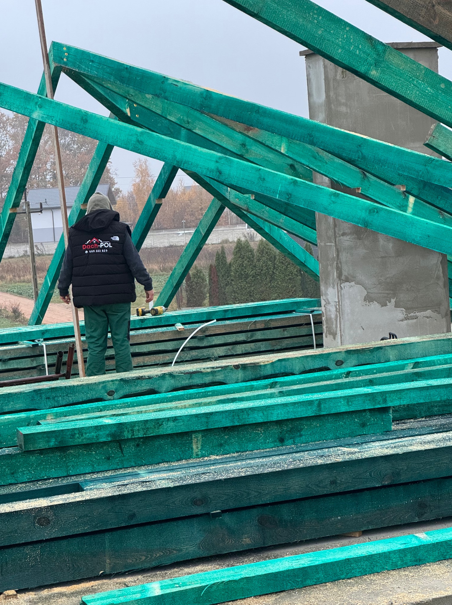 Monter w czarnej kamizelce firmy Dach-Pol montuje zieloną więźbę dachową; w tle komin i zabudowa jednorodzinna.