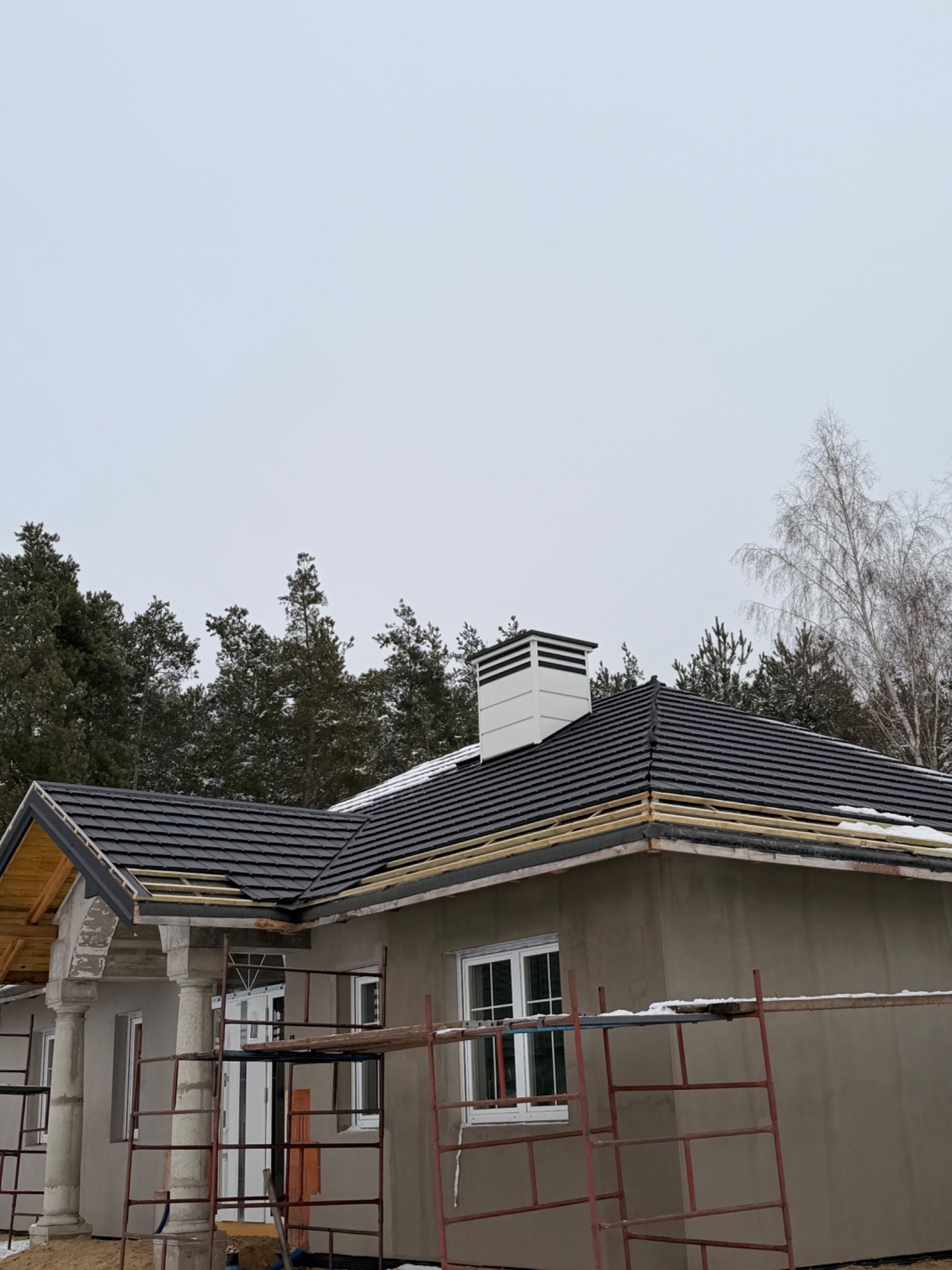 Dom w trakcie budowy dachu z czarną dachówką, widoczne rusztowania i komin. W tle las i pochmurne niebo. Częściowo wykończona elewacja budynku.