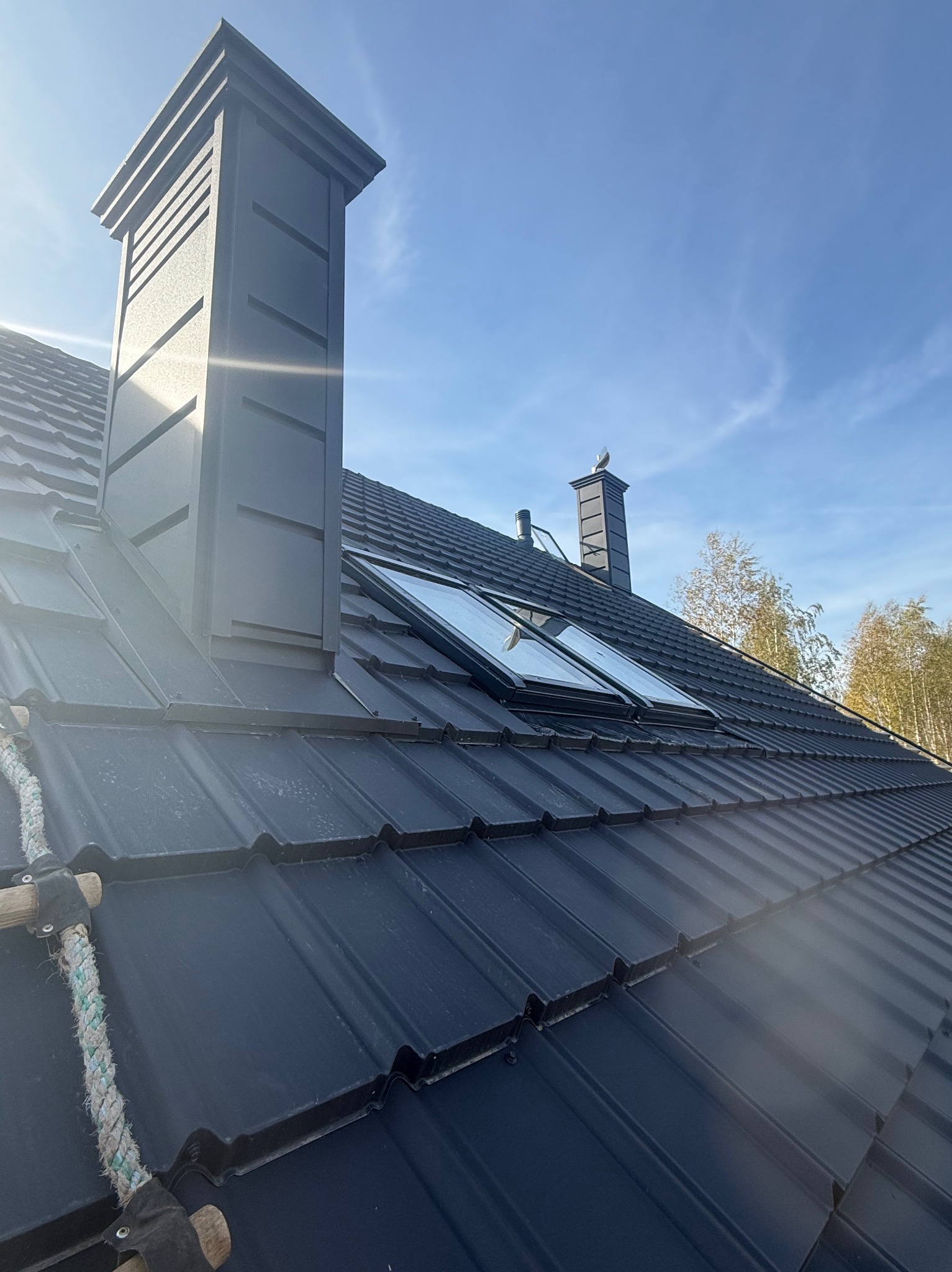 Czarny dach z blachy trapezowej z dwoma oknami dachowymi i kominem z dekoracyjnymi żłobieniami, widziany pod kątem w słoneczny dzień.
