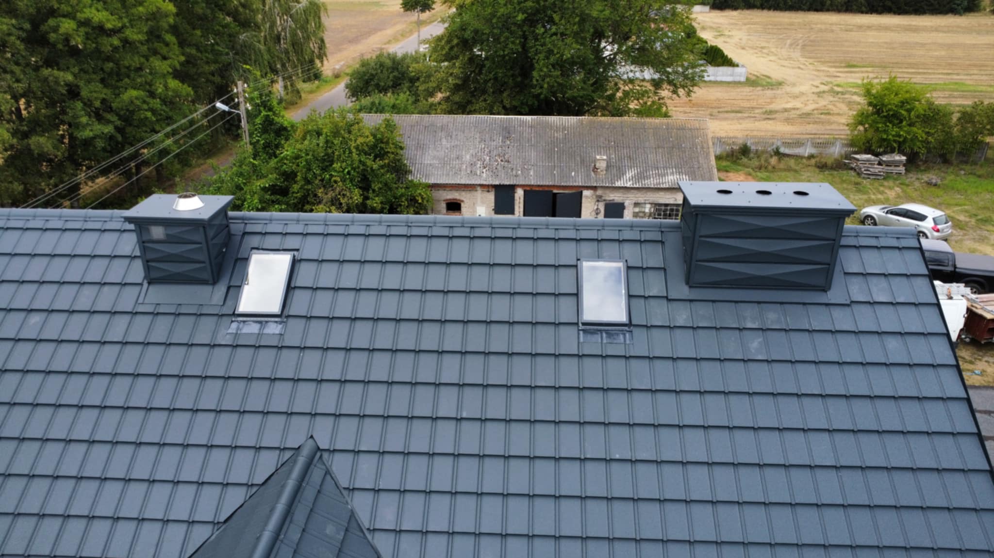 Szary dach z dachówki ceramicznej z dwoma oknami dachowymi i kominami wentylacyjnymi. W tle budynek gospodarczy i pole.