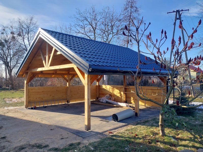 Drewniana altana z niebieskim dachem z blachy trapezowej, widoczna w słoneczny dzień. Na podłodze leżą zwinięte materiały budowlane.