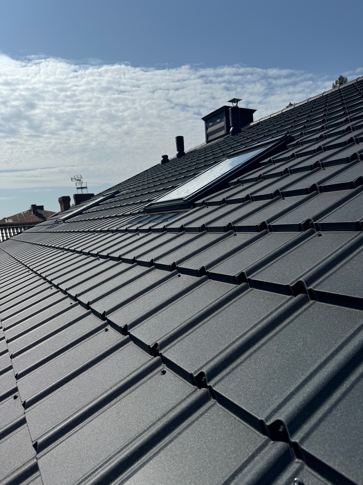 Grafitowy dach z blachodachówki modułowej, z oknem dachowym i kominami na tle błękitnego nieba z chmurami. Widok z dołu, podkreślający regularność wzoru.