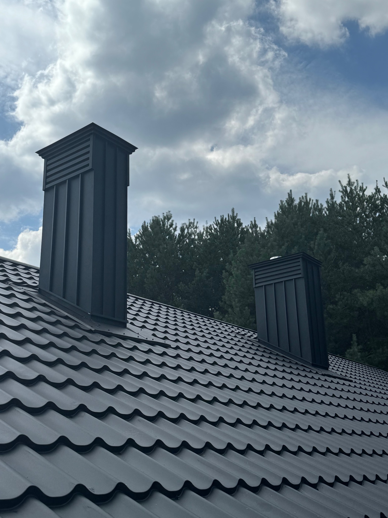 Czarny dach z blachy trapezowej z dwoma kominami wentylacyjnymi na tle pochmurnego nieba i drzew. Widok z dołu, podkreślający geometrię dachu.