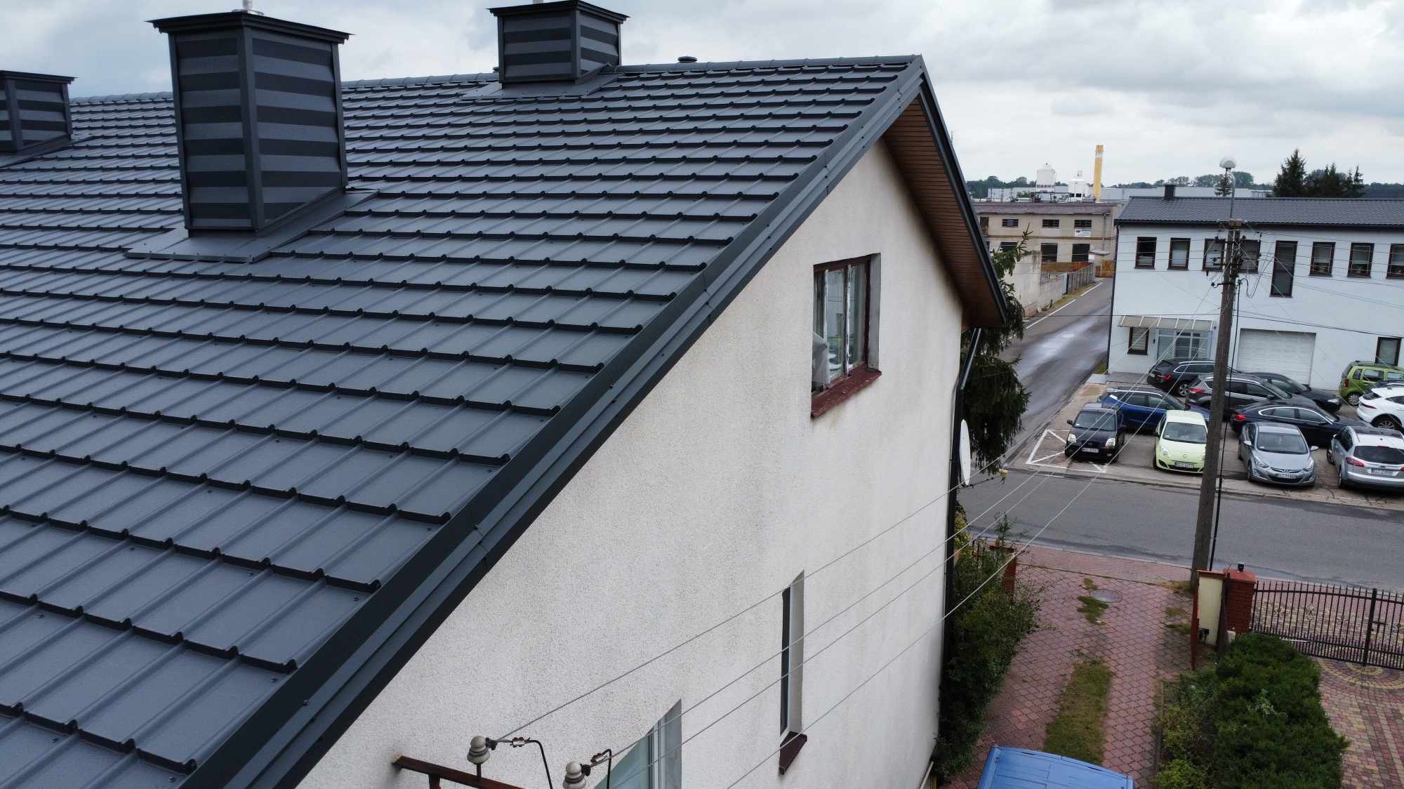 Szary dach z blachy trapezowej z trzema kominami na domu z elewacją w kolorze złamanej bieli. Widok z góry, w tle zabudowa miejska i pochmurne niebo.