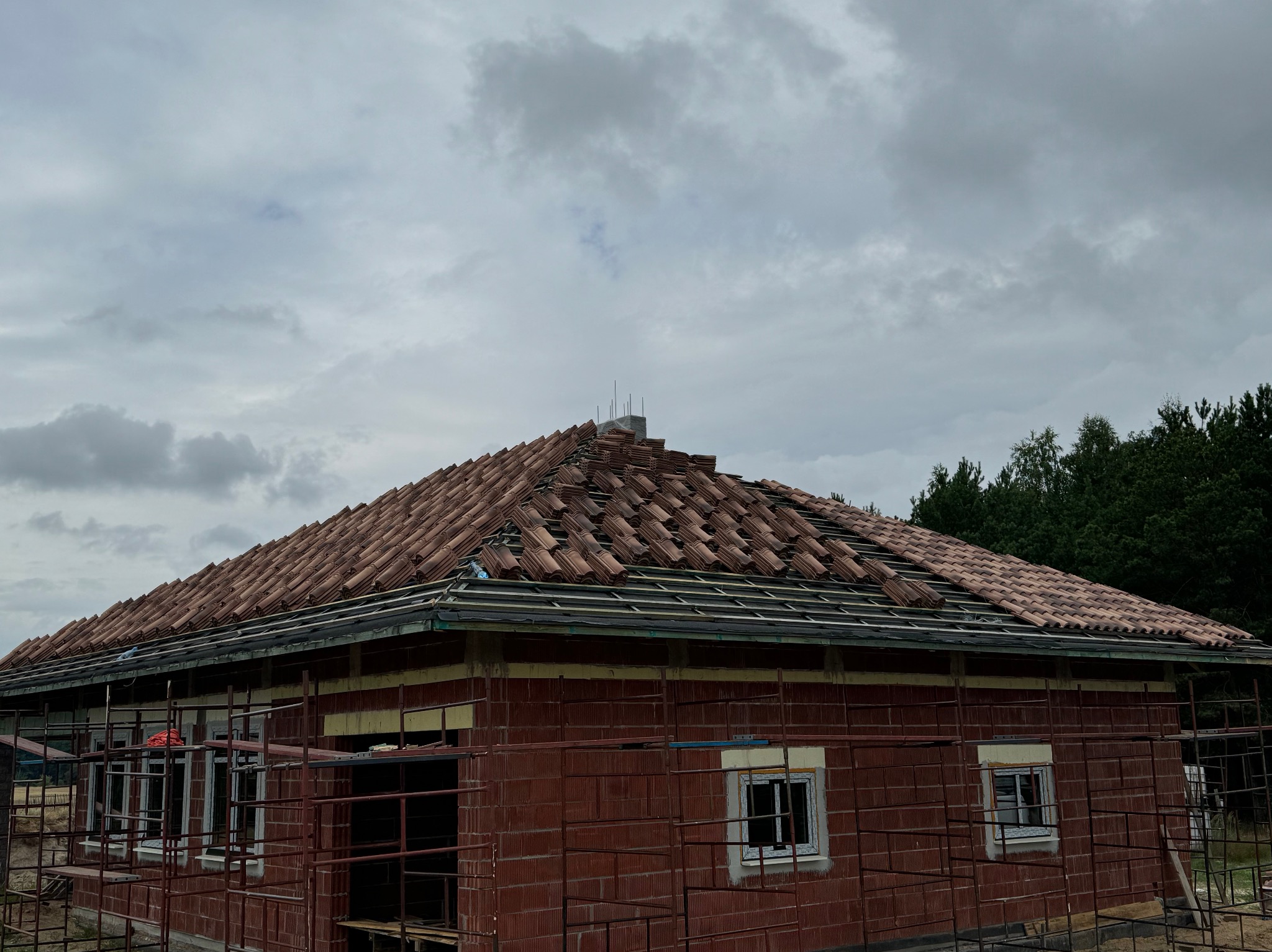 Ceglany dom w trakcie budowy dachu z czerwonej dachówki ceramicznej. Widoczne rusztowania i elementy konstrukcyjne dachu. Pochmurne niebo w tle.