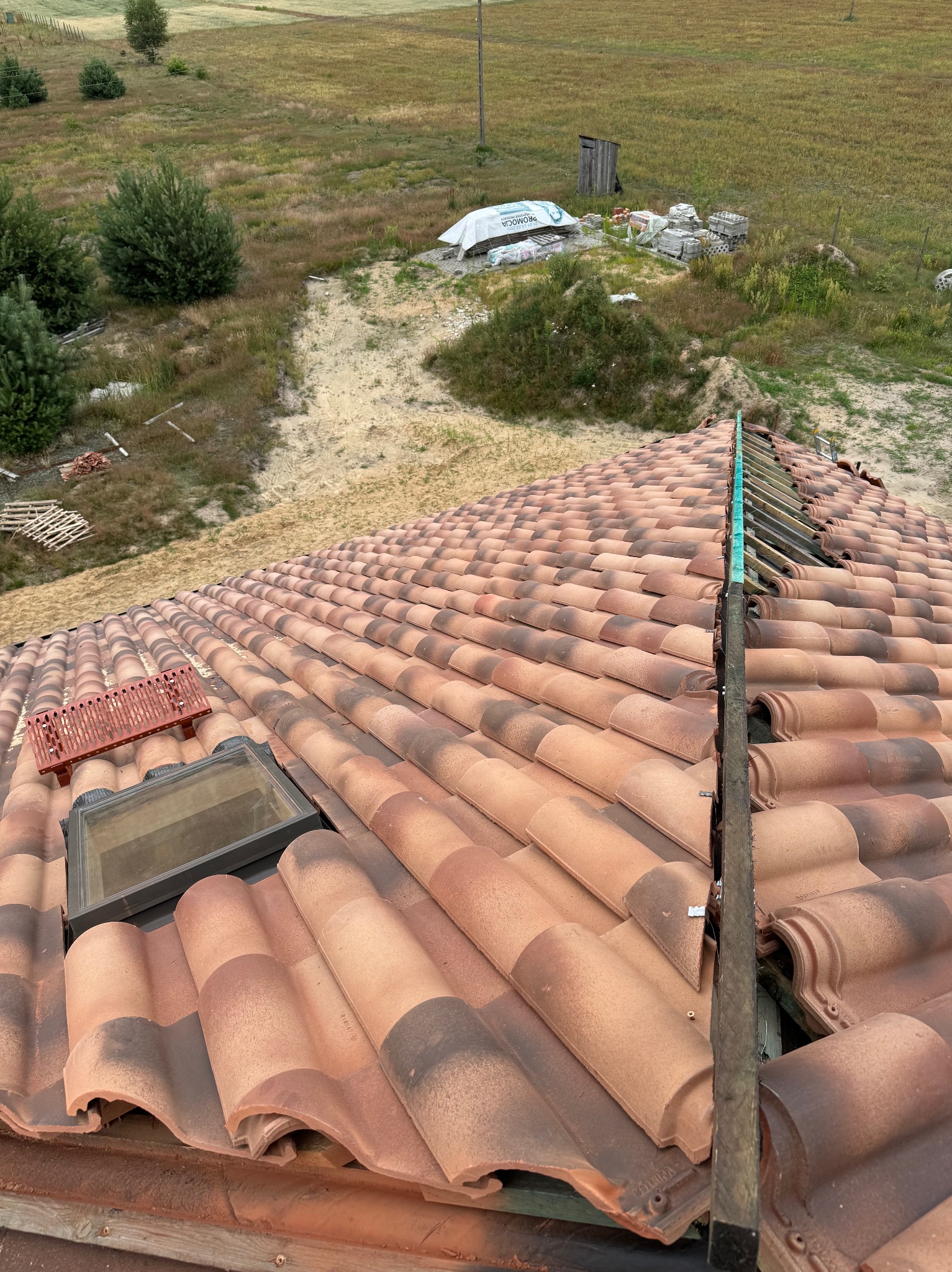 Ułożona dachówka ceramiczna w odcieniach czerwieni i brązu, z widocznym oknem dachowym i elementami montażowymi. W tle krajobraz z polami i drzewami.