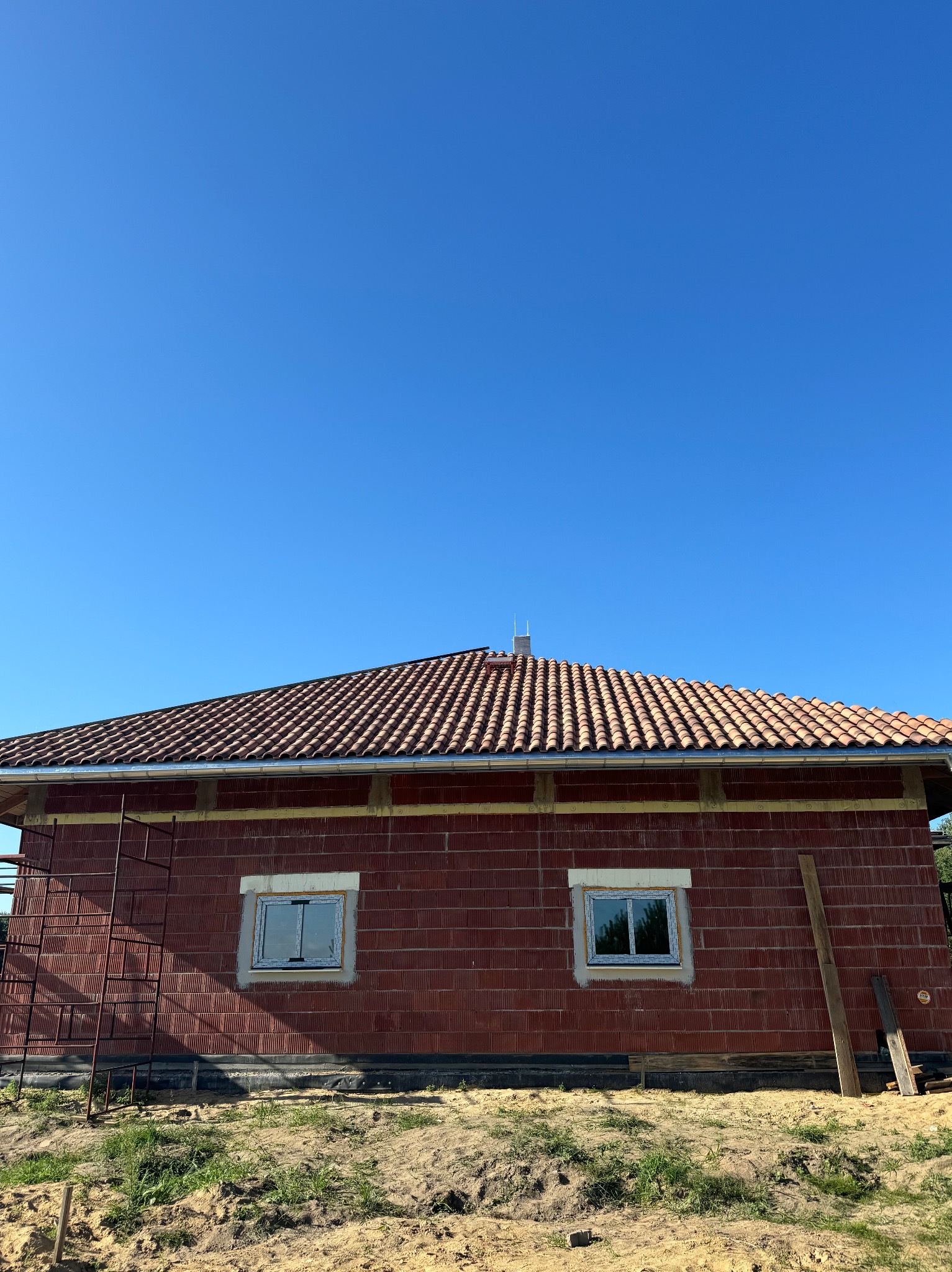 Dom w trakcie budowy z czerwoną dachówką i oknami. Widoczne rusztowanie i elementy konstrukcyjne. Błękitne niebo w tle.