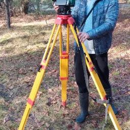 Mężczyzna w kurtce i czapce ustawia teodolit na żółtym statywie w lesie, widoczne opadłe liście.
