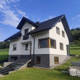 USŁUGI BUDOWLANE MAREK ŻĄDŁO - Dwukondygnacyjny dom z ciemną dachówką i jasną elewacją w Jordanowie, z szarym cokołem i schodami wejściowymi, na tle zielonego wzgórza i błękitnego nieba.