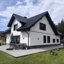USŁUGI BUDOWLANE MAREK ŻĄDŁO - Biały dom z czarnym dachem i obramowaniami okien, otoczony zielonym trawnikiem i szarą kostką brukową. Widoczne meble ogrodowe i zabawki.