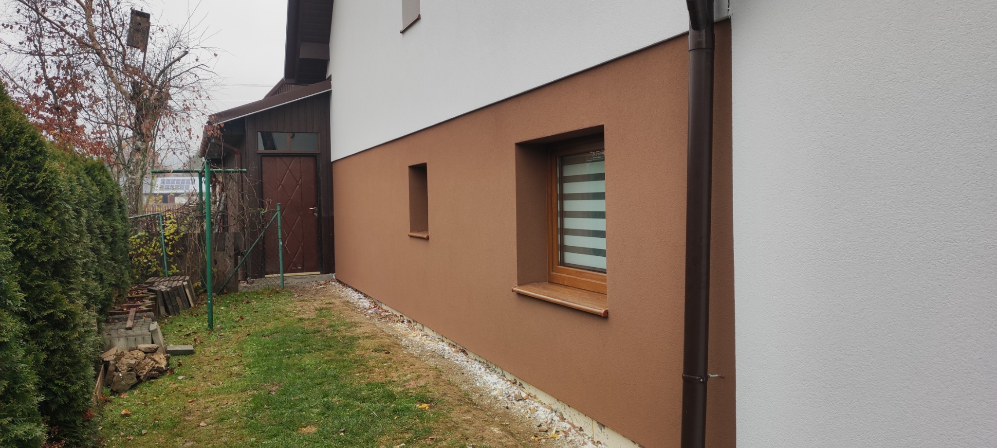 Dwukolorowa elewacja domu z oknami z drewnianą ramą, brązowy tynk na dolnej części ściany i biały na górnej, widoczny fragment rynny i niewielki fragment ogrodu.