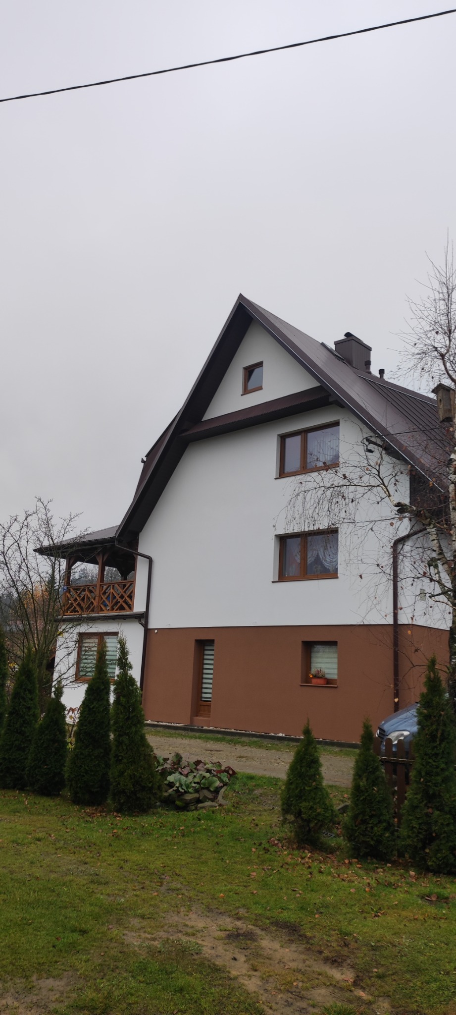 Dwukondygnacyjny dom w Jordanowie z białą elewacją i brązowym cokołem, widoczny z zewnątrz, z brązowymi oknami i dachem, otoczony zielonymi tujami i trawnikiem w pochmurny dzień.