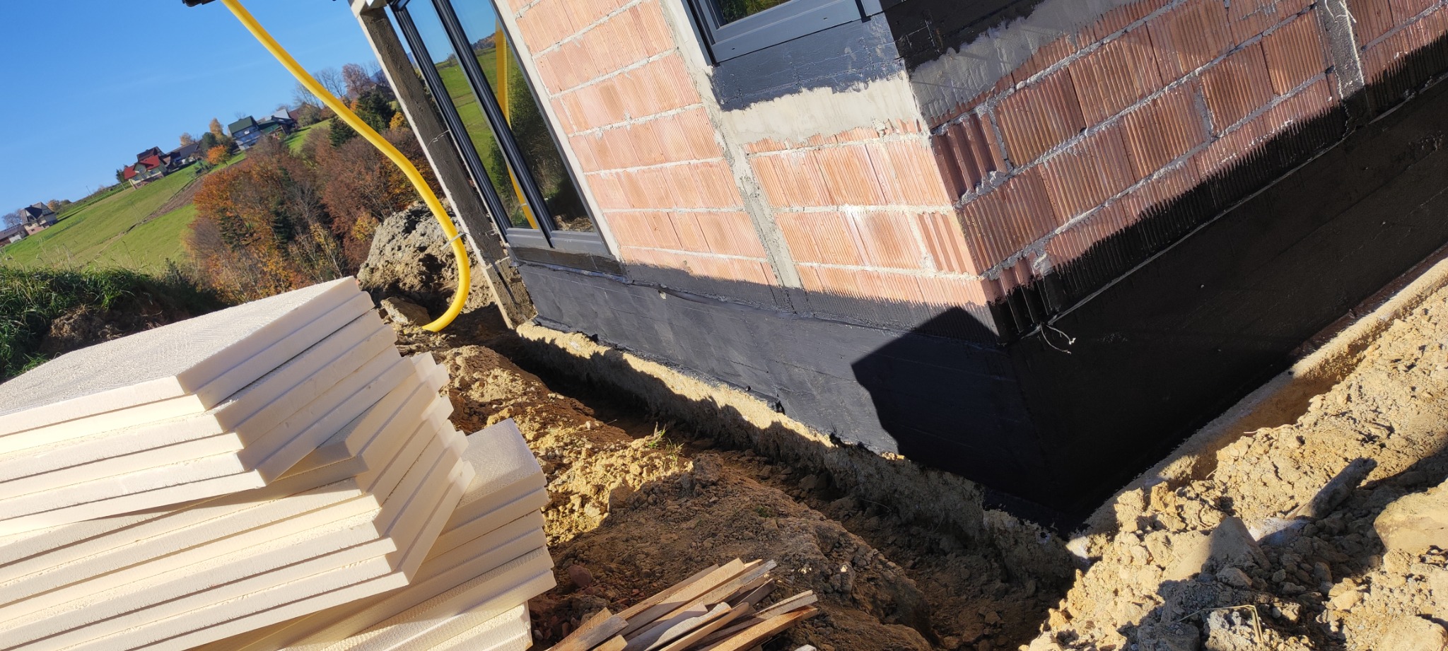 Fundament budynku z oknem, zabezpieczony czarną izolacją, obok stos białych płyt izolacyjnych i wykopana ziemia.