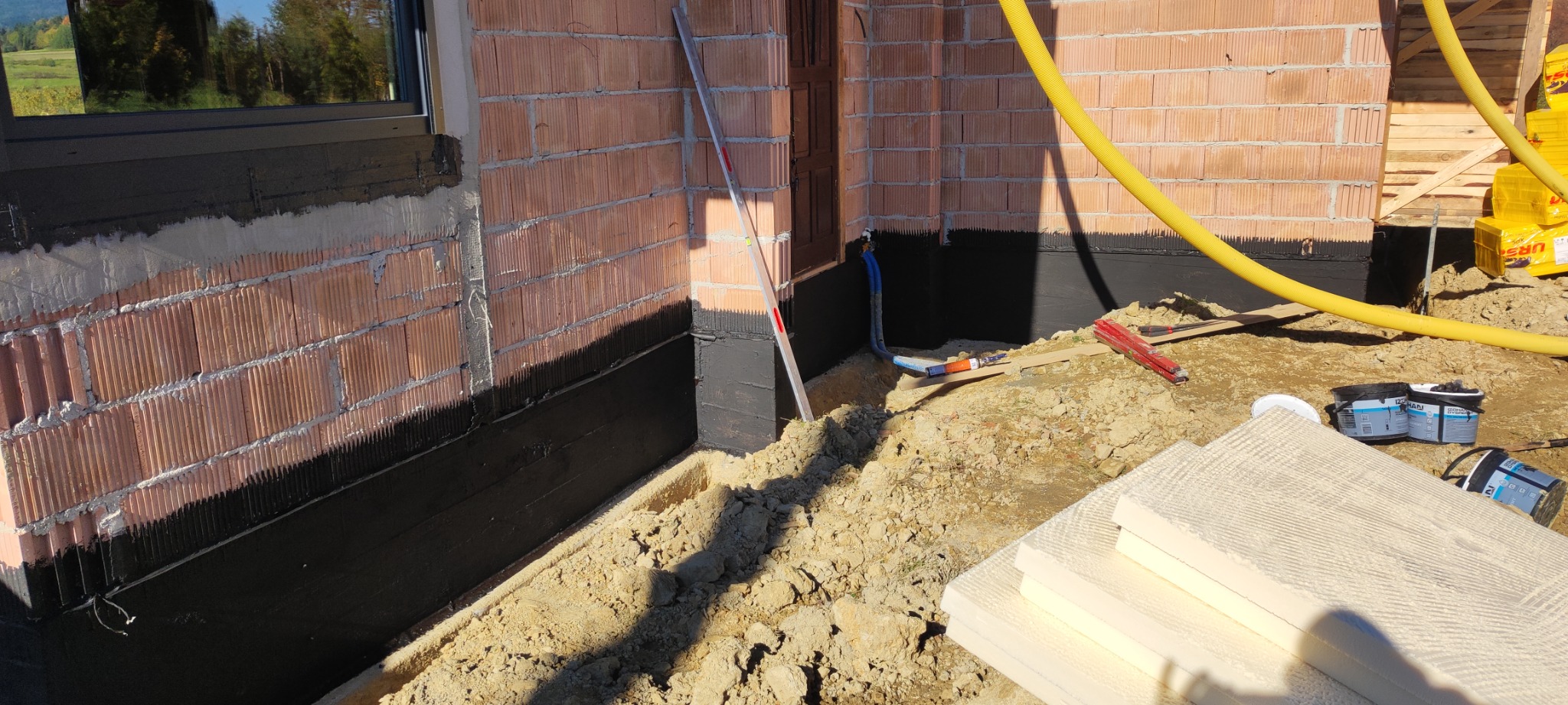 Fundament budynku w trakcie izolacji, widoczna warstwa hydroizolacji, styropian do ocieplenia, poziomica oparta o ścianę, żółty wąż i pojemniki z masą uszczelniającą.