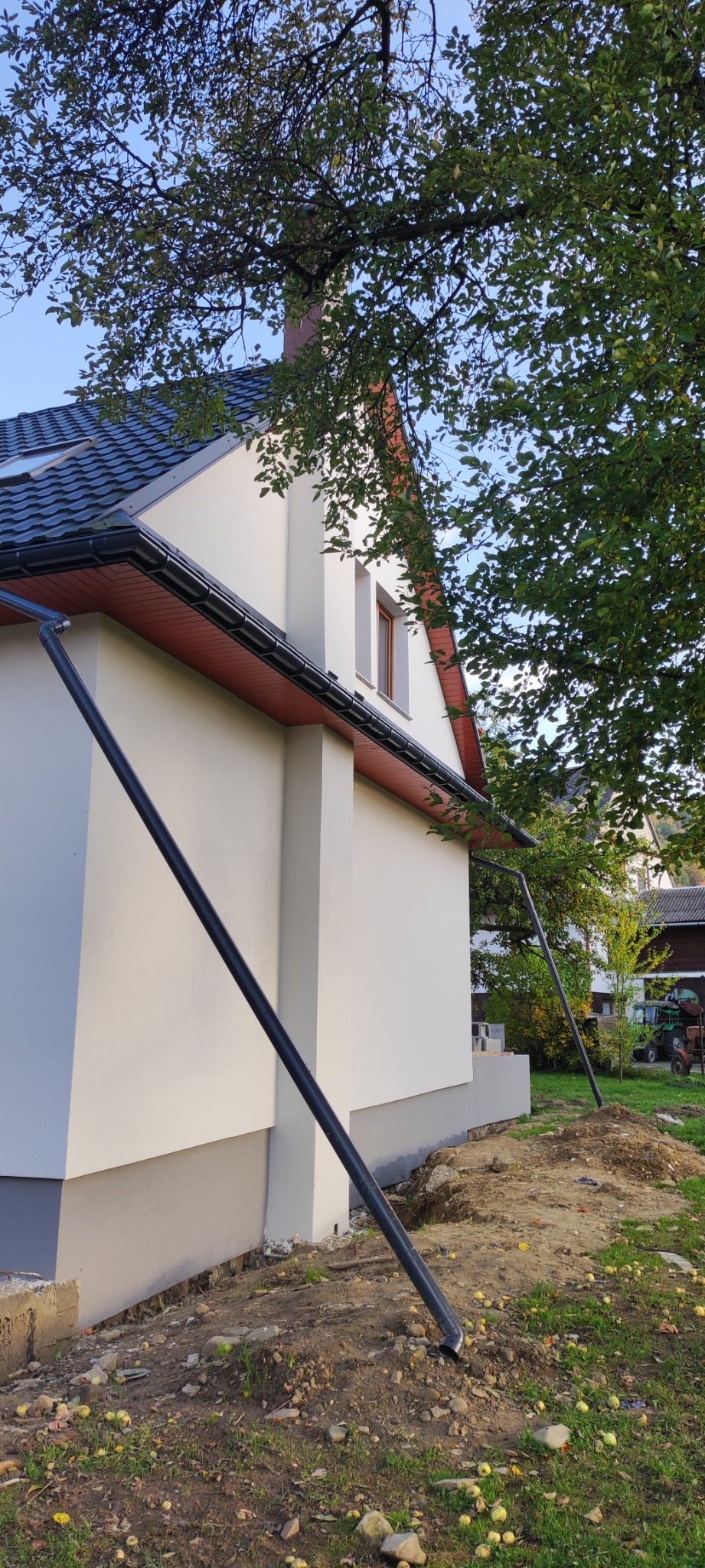 Boczny widok ocieplonego domu jednorodzinnego z grafitową podmurówką, nową elewacją i systemem odprowadzania wody deszczowej, widoczne jabłka na ziemi.