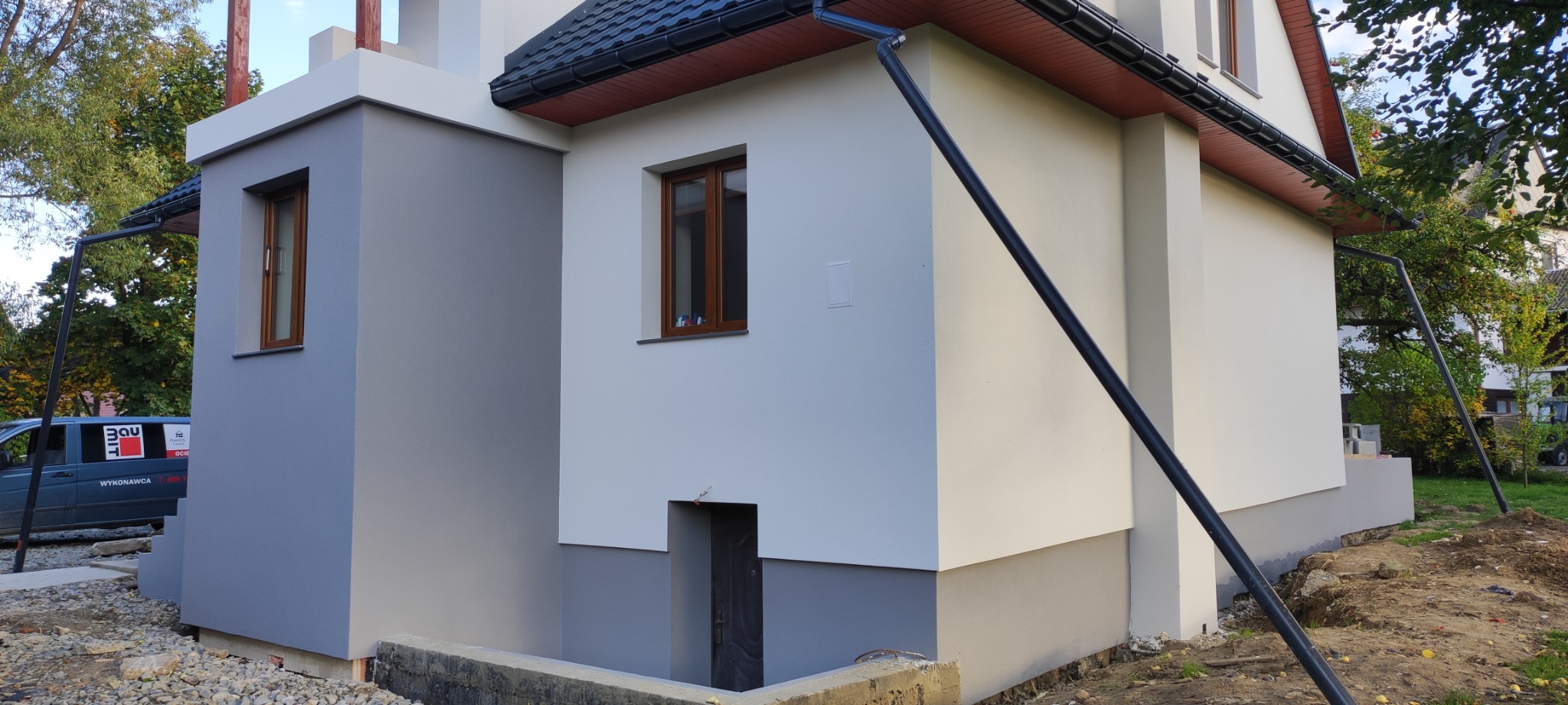 Dwukondygnacyjny budynek po tynkowaniu elewacji, z widocznym fragmentem fundamentu pomalowanego na szaro, brązowe ramy okienne i czarna rura spustowa, na tle zieleni i firmowego samochodu.