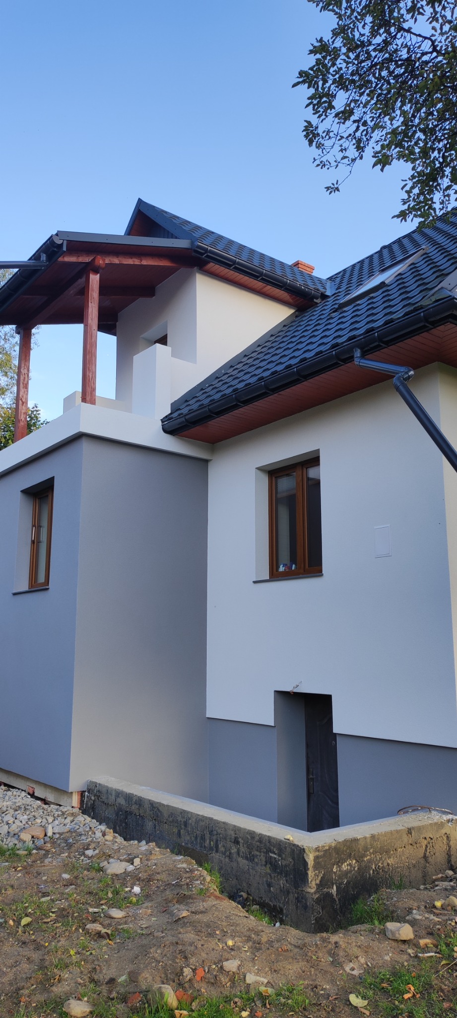 Dwukondygnacyjny dom z elewacją w odcieniach szarości i bieli, z widocznymi detalami wykończenia, takimi jak okna z drewnianymi ramami, czarny dach z rynnami i betonowe fundamenty w ziemi.