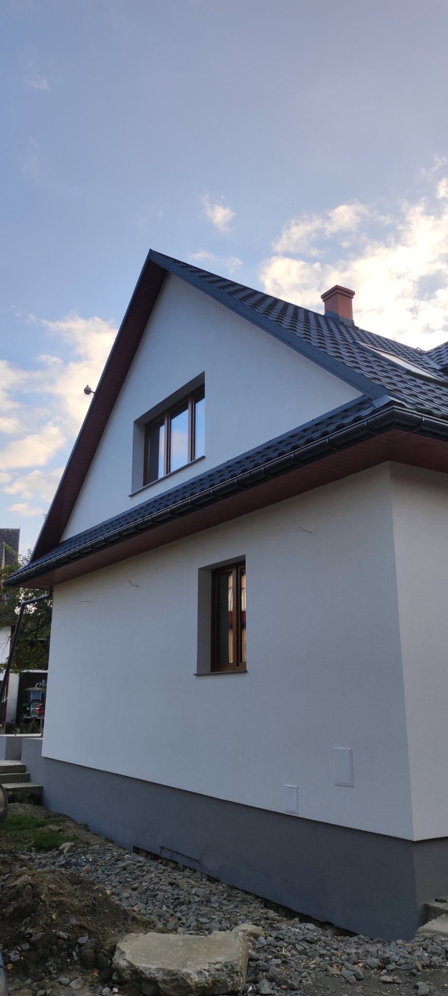Wykończona elewacja domu z dwoma oknami o brązowych ramach, ciemnym dachem i świeżo pomalowanymi fundamentami, widok z boku na tle błękitnego nieba.
