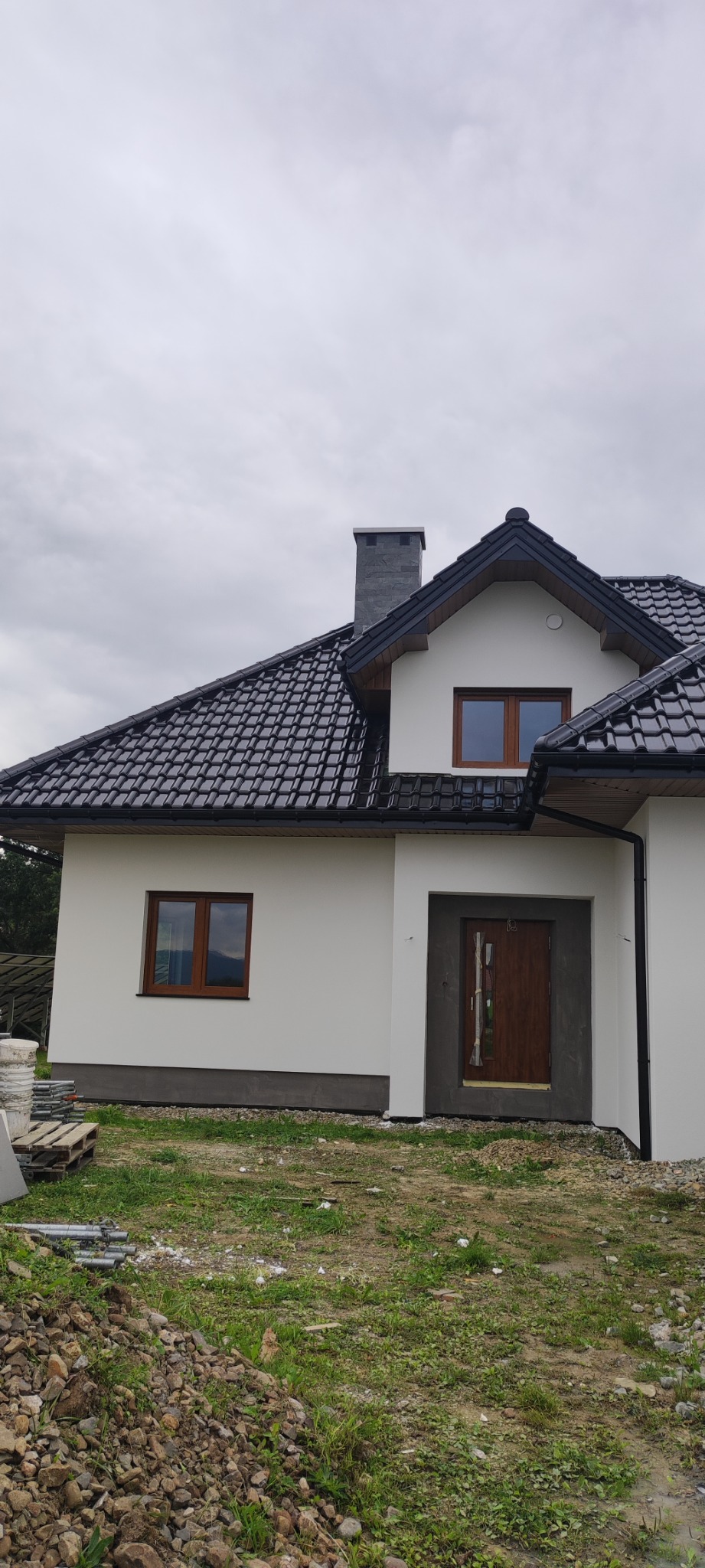 Wykończona elewacja domu jednorodzinnego z ciemną dachówką i stolarką okienną w odcieniu drewna, widoczne palety i gruz po budowie.