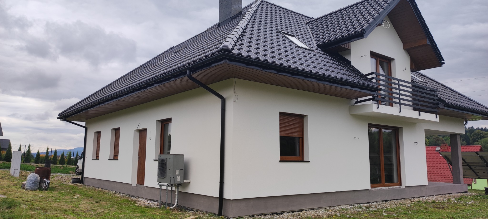 Świeżo otynkowany dom jednorodzinny z ciemną dachówką i drewnianą stolarką okienną, widoczny z zewnątrz, z zamontowanym klimatyzatorem przy ścianie.