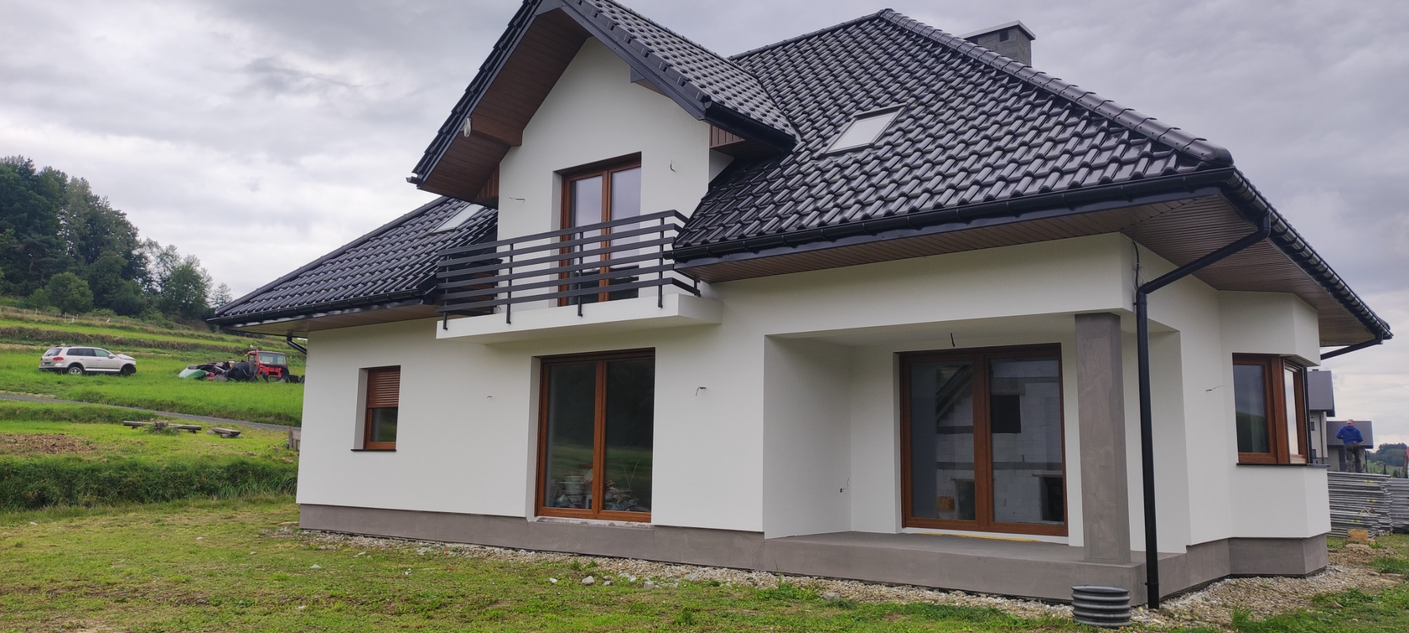 Nowoczesny dom jednorodzinny z białymi ścianami i ciemną dachówką, z balkonem i dużymi oknami, widok z zewnątrz na tle pagórkowatego terenu z pojazdami.
