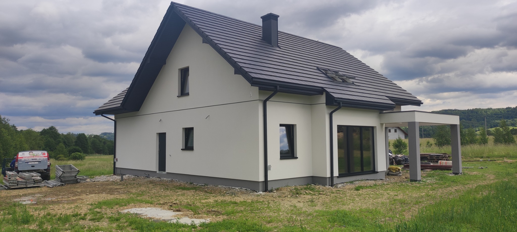Nowoczesny dom jednorodzinny z jasną elewacją i ciemną dachówką, widok z boku na tle zachmurzonego nieba i zieleni, z widocznym autem firmy budowlanej w tle.