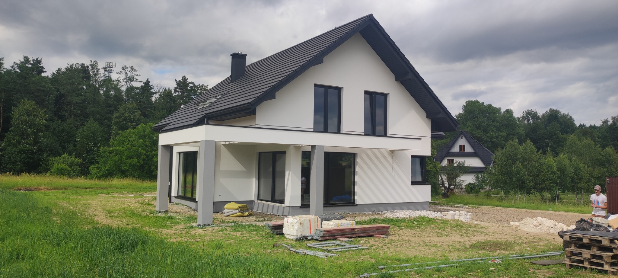 Nowoczesny dom jednorodzinny z białymi ścianami i czarnym dachem, widoczny z zewnątrz w pochmurny dzień, z elementami przygotowanymi do dalszych prac wykończeniowych wokół budynku.